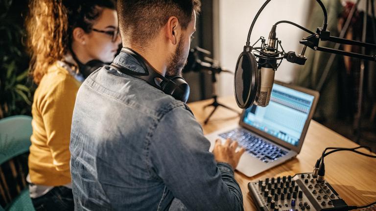 A man and a woman with headphones on sitting in front of a laptop and recording equipment