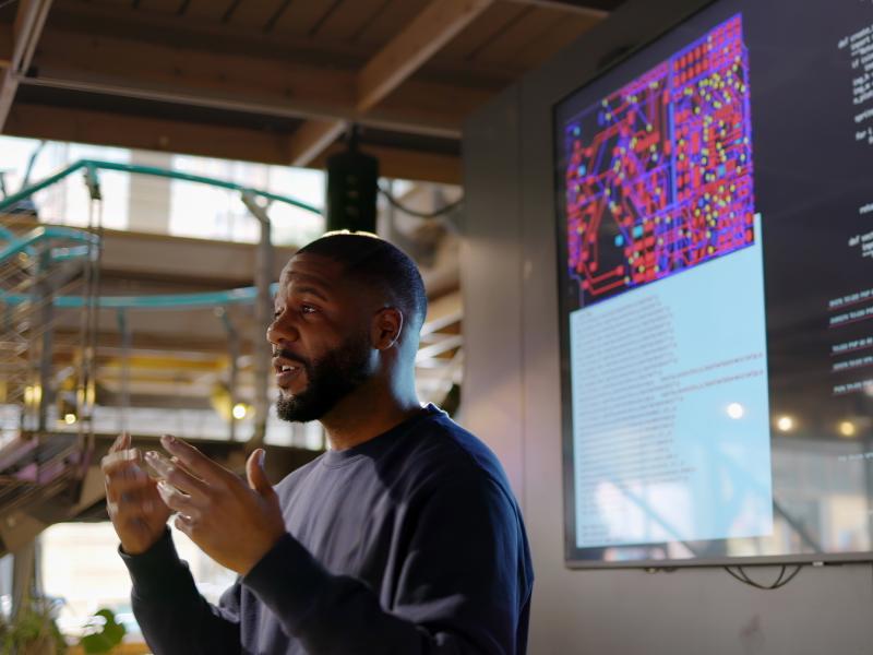 African American man standing on a stage addressing an audience, with coding information on a screen behind him